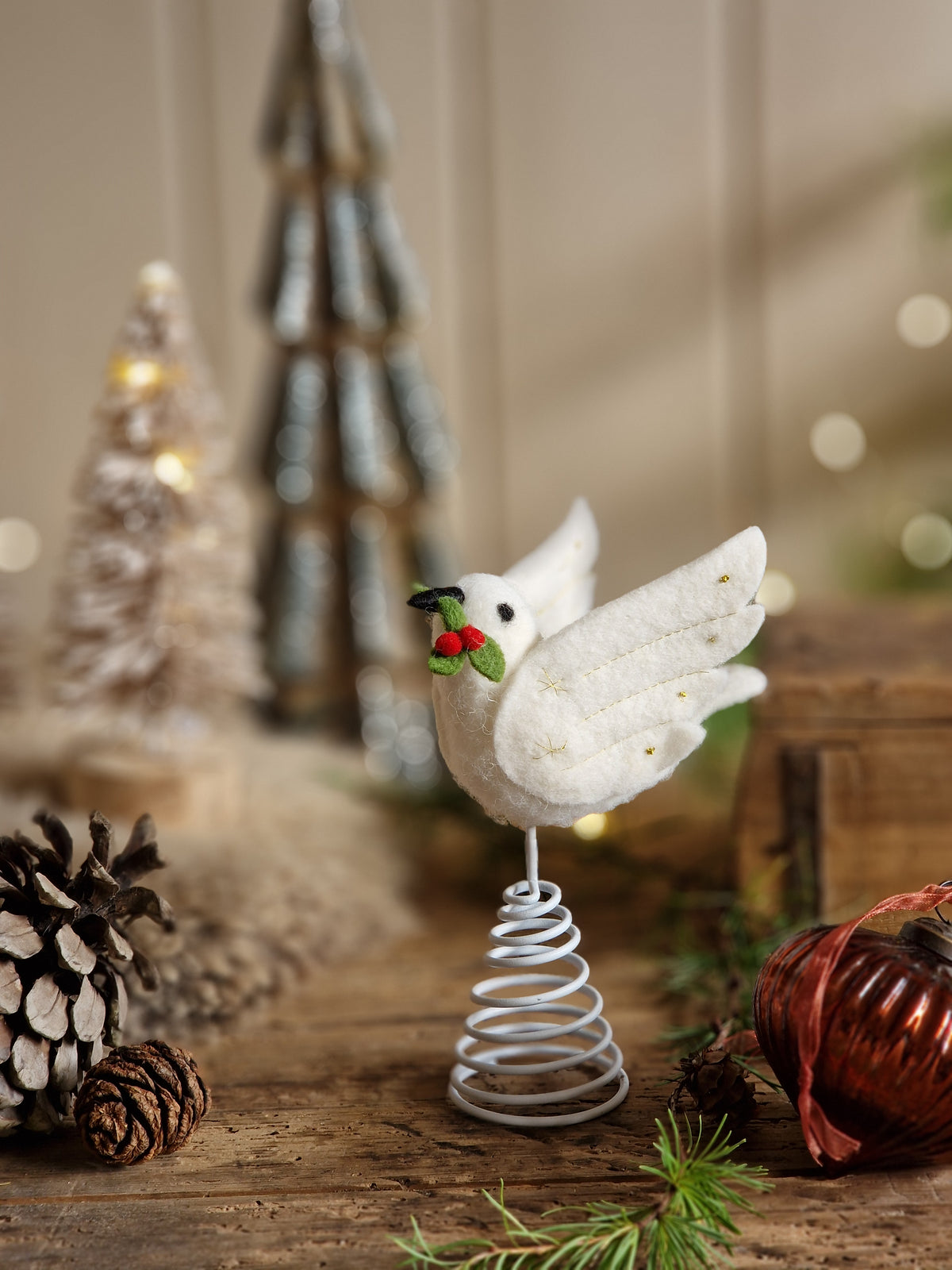 A white felt Christmas dove ornament with a small green leaf and red berry in its beak, mounted on a silver spiral spring stand, displayed on a rustic wooden surface with pinecones and holiday decor in the background.