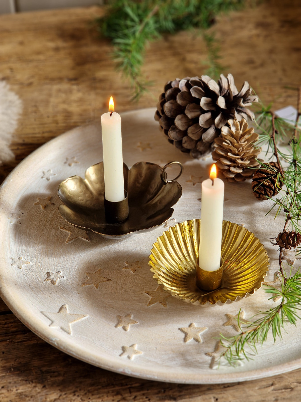 A decorative ceramic Advent wreath holder with three white taper candles, styled with pinecones and evergreen branches on a rustic wooden surface.