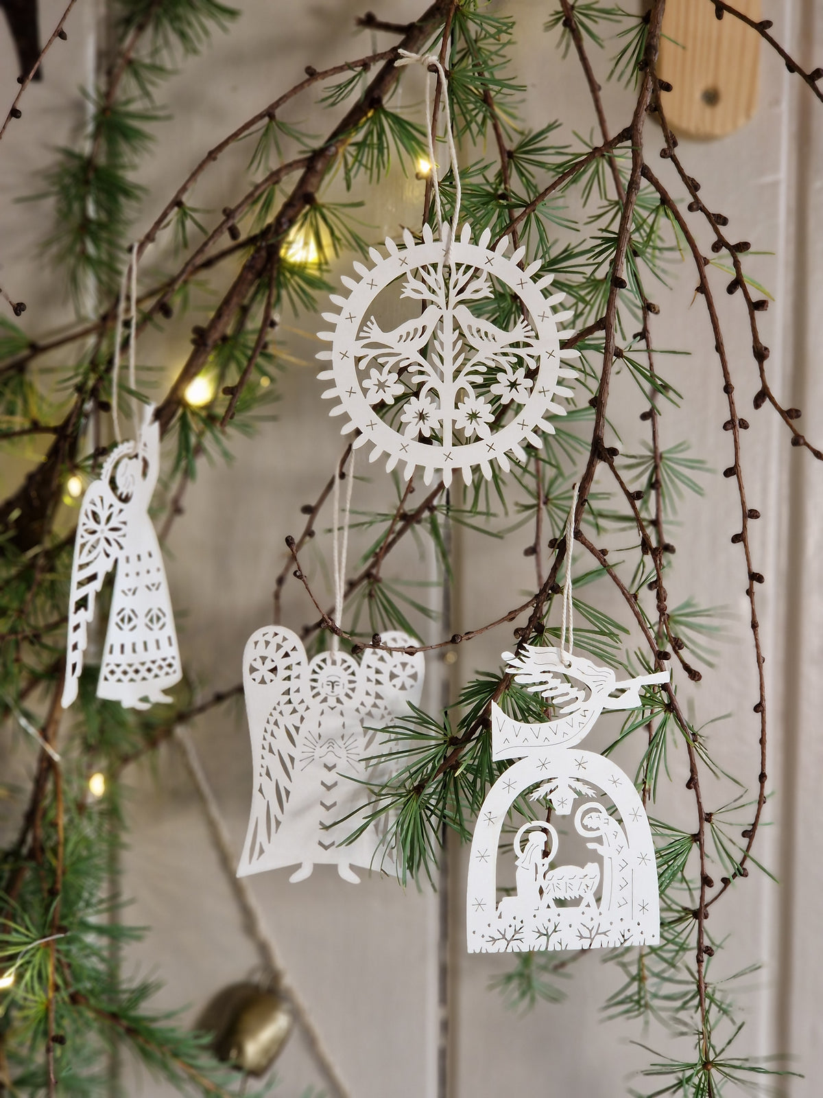 White paper cutout Christmas ornaments hanging from bare tree branches with green pine needles and warm string lights in the background.