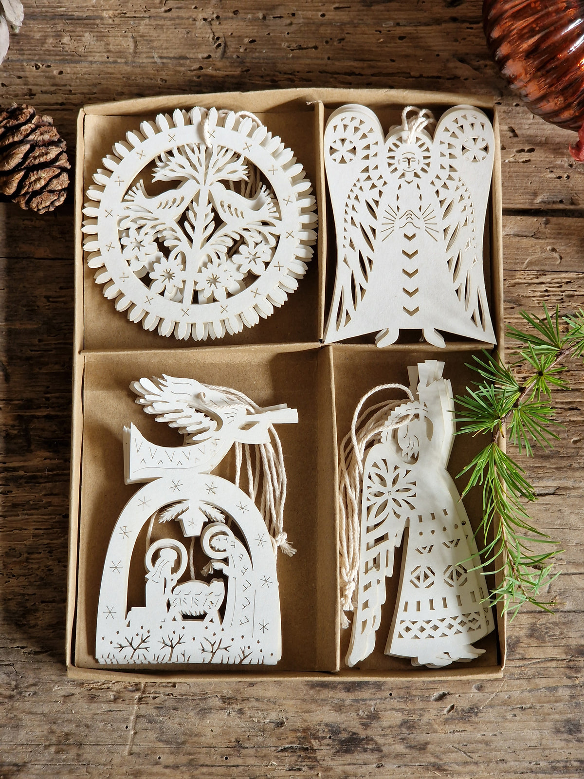 Four white laser-cut wooden Christmas ornaments displayed in a brown cardboard tray on a rustic wooden table, surrounded by pine branches, a pinecone, and holiday decor.