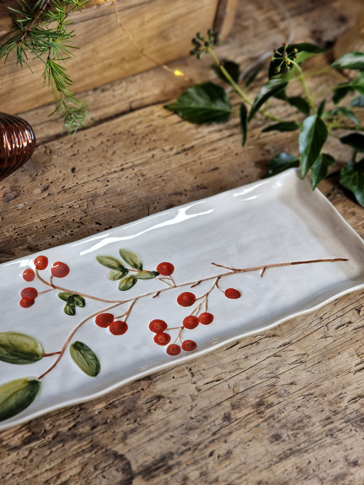 A rectangular white ceramic serving platter decorated with a painted holly branch motif featuring green leaves and red berries, styled on a rustic wooden tabletop with green foliage.