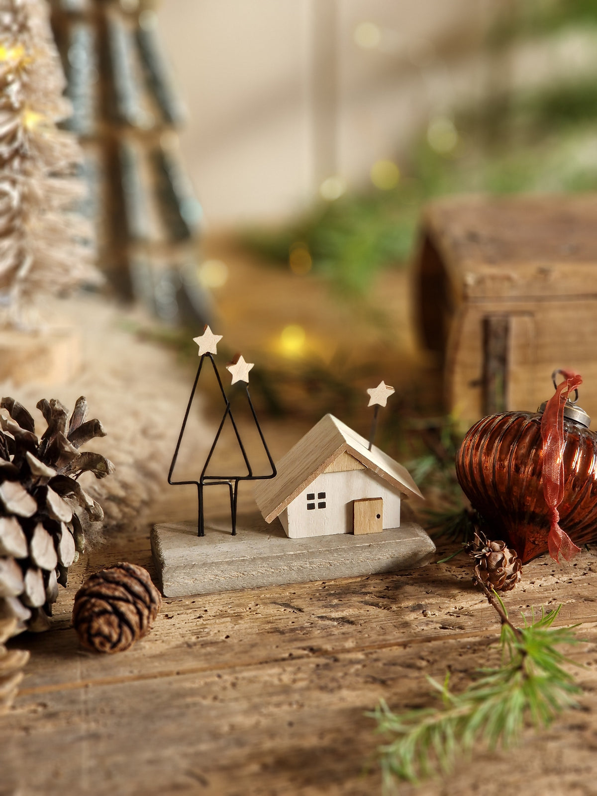 A close-up of rustic holiday decorations on a weathered wooden surface, including a small white house ornament, pinecones, evergreen branches, and a copper-colored ornament, with soft warm bokeh lights in the background.