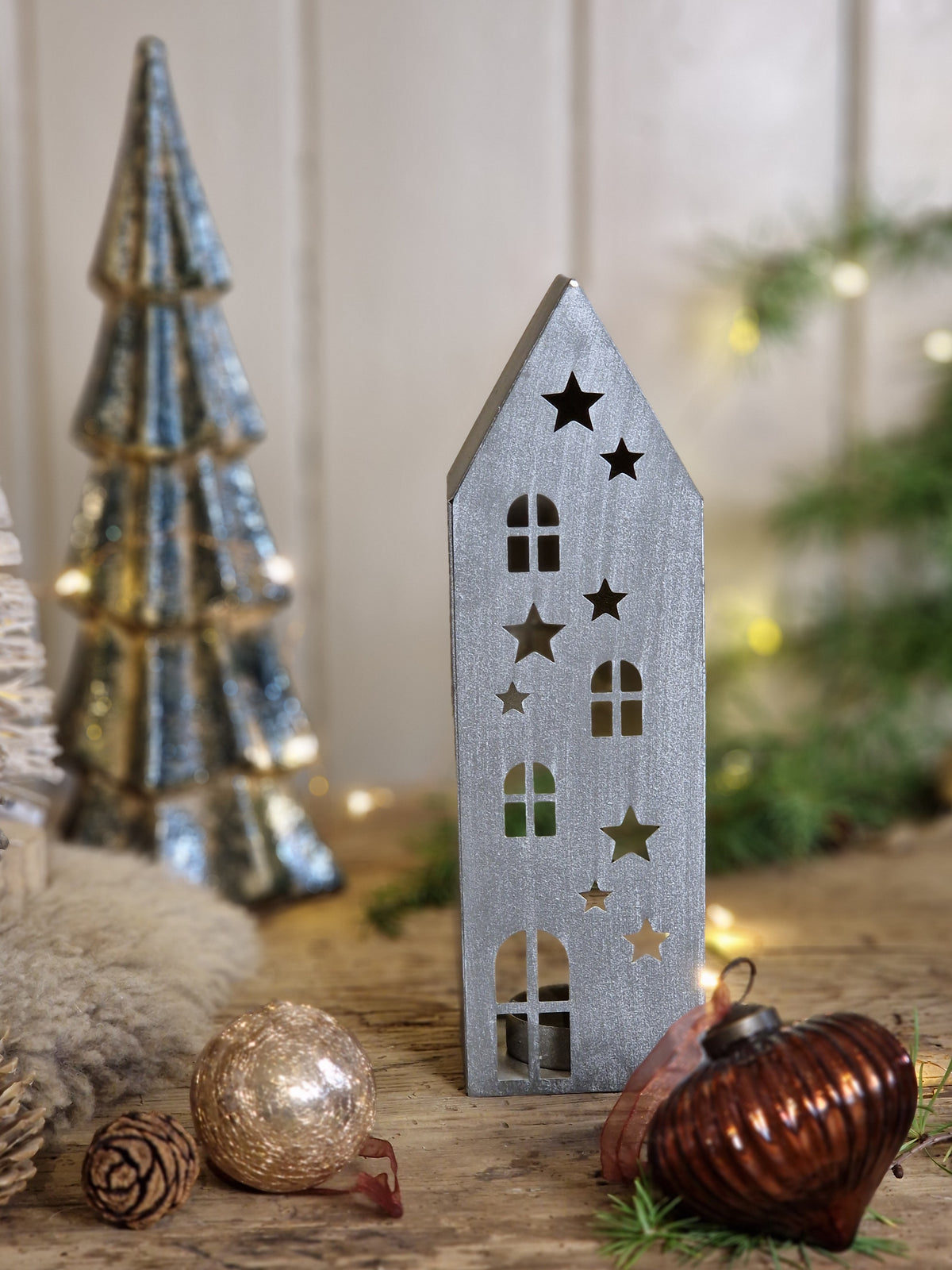 A small silver wooden house ornament with star and window cutouts, displayed on a rustic wooden surface alongside a metallic silver tree ornament, a gold ball ornament, and a brown heart-shaped ornament.