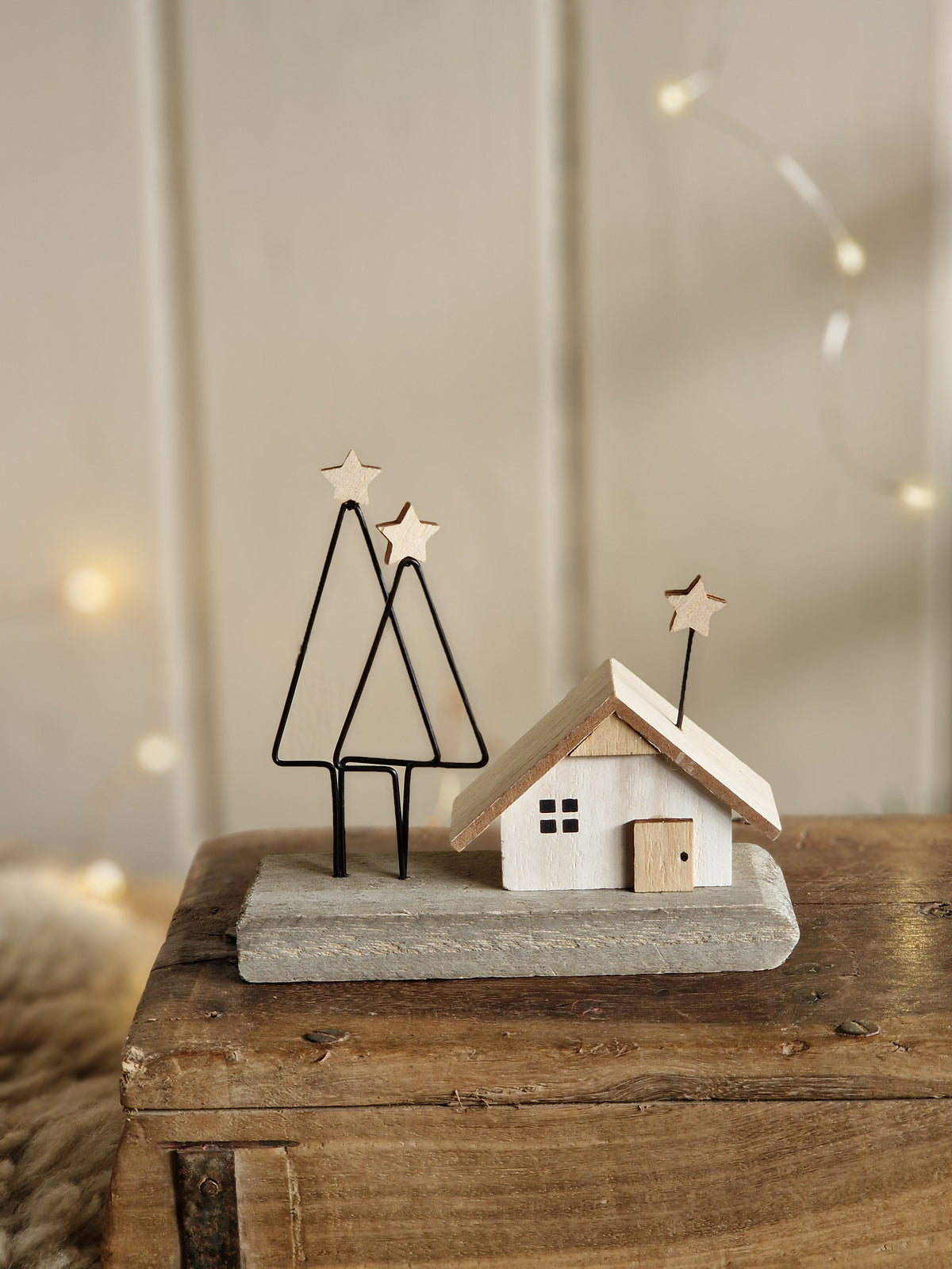 A rustic holiday wall decoration featuring a small white wooden house with a gray stone-textured base, mounted on a wooden panel with wire tree silhouettes and star accents.