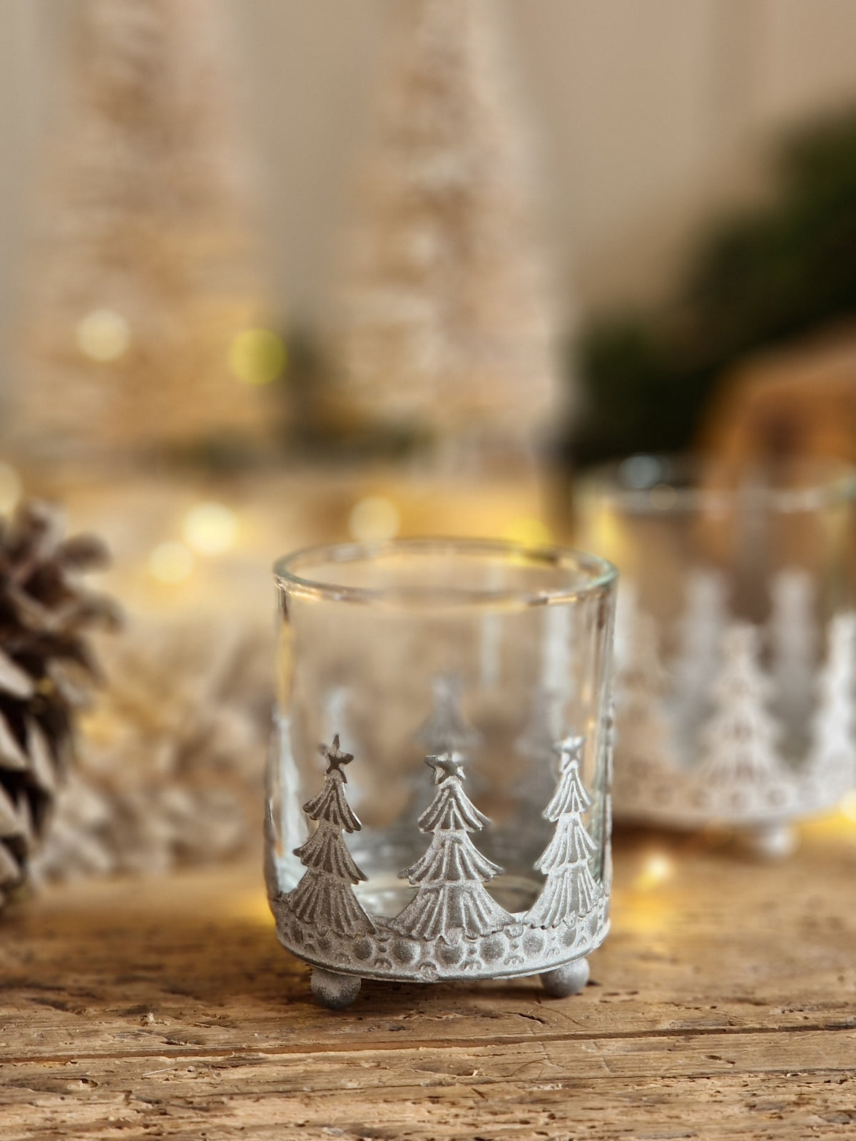 A clear glass candle holder with a white-painted decorative border of three Christmas trees, shown on a rustic wooden surface with warm bokeh lights in the background.