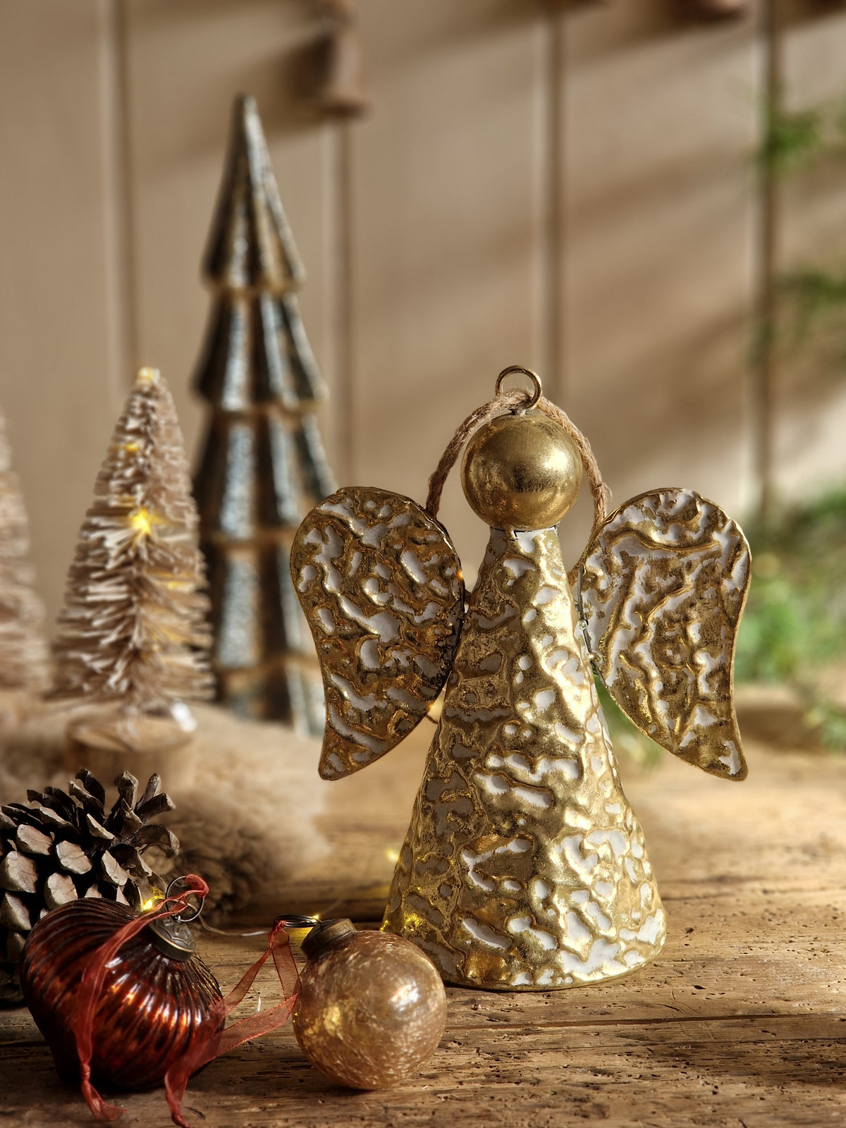 A textured metallic gold Christmas angel ornament standing on a rustic wooden surface, with small decorative trees, a pinecone, and a red ornament in the foreground against a softly blurred beige background.