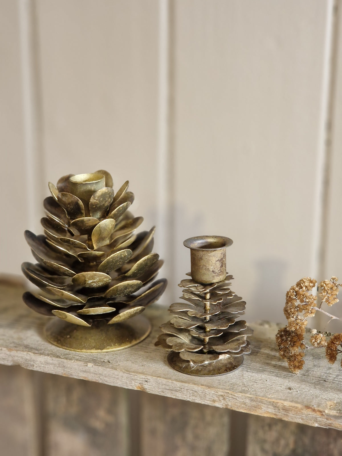 Two vintage-style pinecone-shaped candle holders in aged brass and silver-toned metal, displayed on a rustic wooden shelf against a light paneled wall.