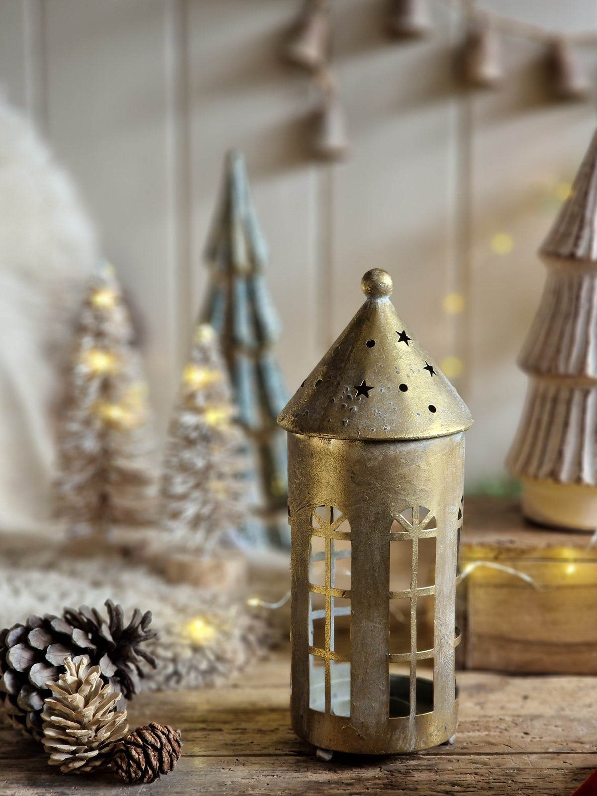 A rustic brass holiday lantern with star cutouts on its pointed roof, styled on a wooden surface with pinecones and miniature Christmas trees in the background.