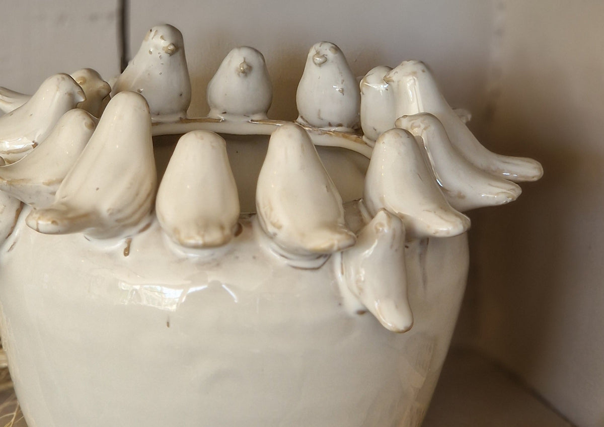 A handmade ceramic pot with a raised rim of small bird figures, shown on a rustic shelf next to wooden dominoes.