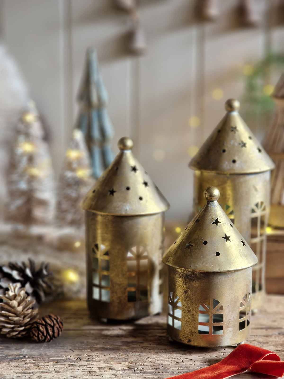 Three small house-shaped brass lanterns with star and window cutouts, arranged on a rustic wooden surface with pinecones and warm blurred holiday decor in the background.