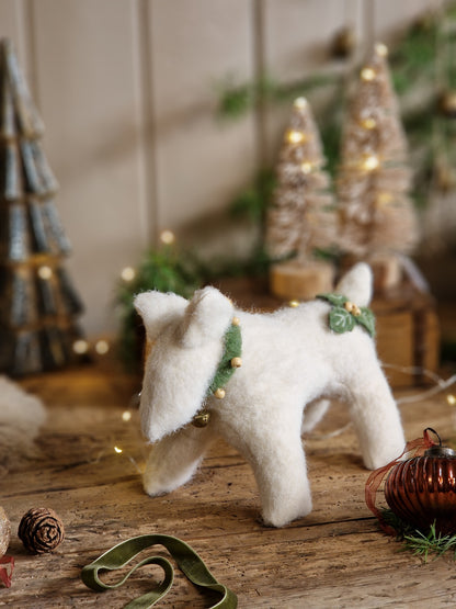 A white felt Christmas dog ornament with a green leaf collar and small gold bells, standing on a rustic wooden surface with blurred holiday decorations in the background.