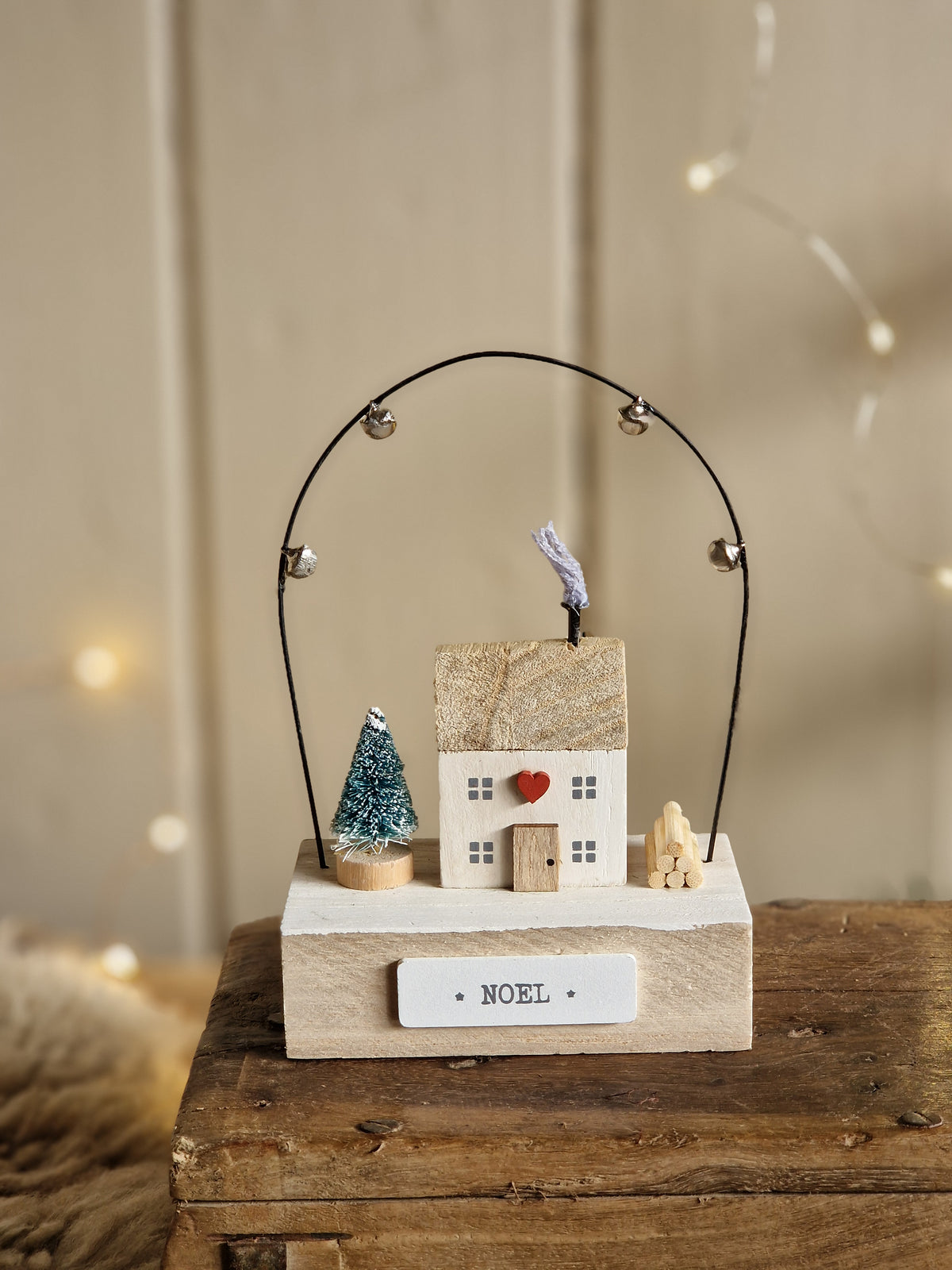 A close-up of a wooden Christmas ornament or decorative hanging piece mounted on a rustic wooden post. The ornament features a white rectangular tag with the word 'NOEL', a small map-shaped card, a red button, a blue textured accent, and a black wire loop with small silver beads.