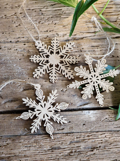 Three laser-cut wooden snowflake ornaments with natural light wood grain and hanging twine, arranged on a rustic weathered wood background with green leaves.