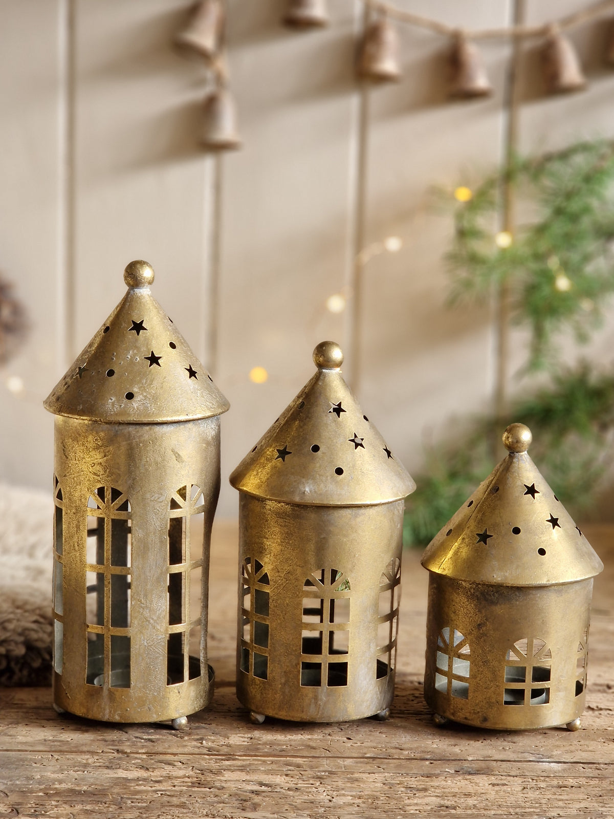 Three small gold-toned metal lanterns with star and window cutouts, arranged on a rustic wooden surface against a softly blurred holiday background.