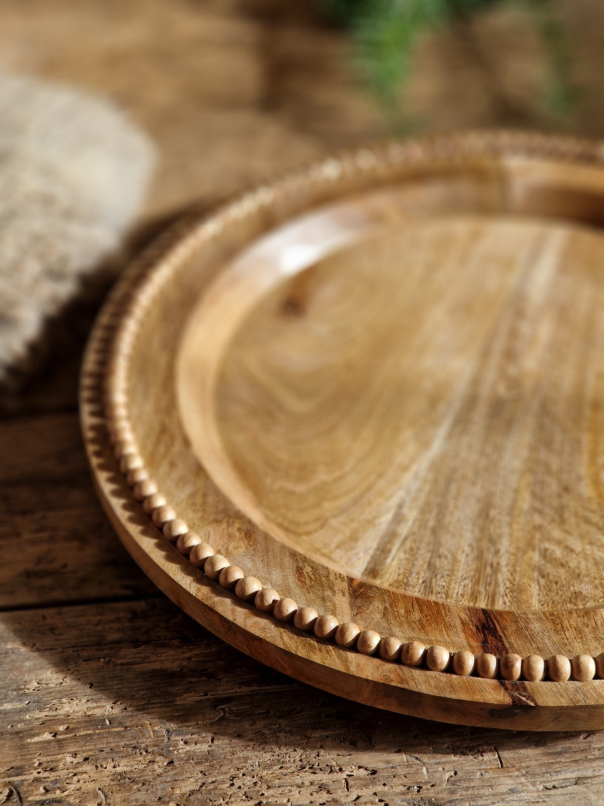 A close-up of a round wooden serving tray with a carved beaded rim, resting against a rustic wooden surface.