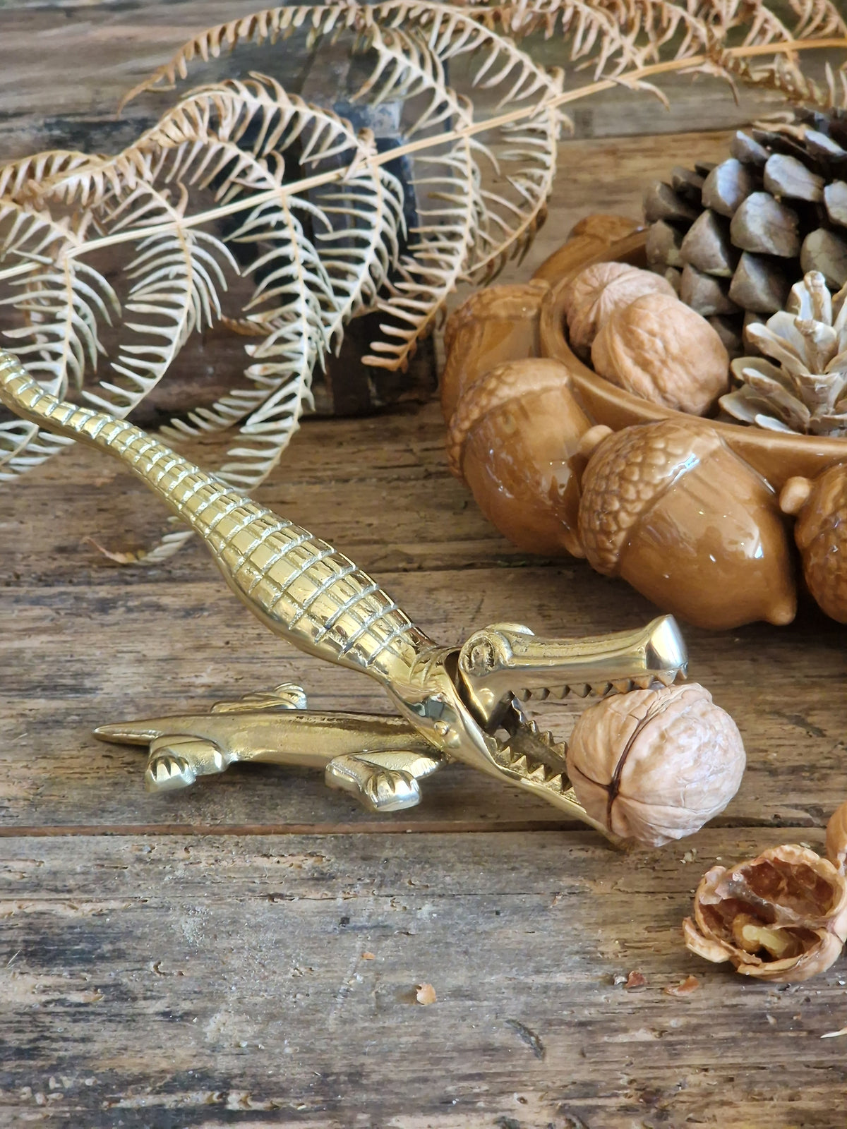 A brass alligator-shaped nutcracker holding a walnut on a rustic wooden surface, styled with acorns, pinecones, dried leaves, and a walnut shell.