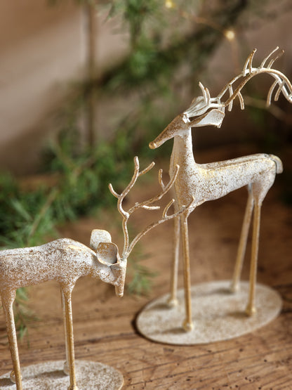 A close-up of a rustic metal deer wall hanging mounted on a wooden wall. The deer figure is textured with a distressed, speckled finish in a light beige or off-white color, with thin metal legs extending horizontally.