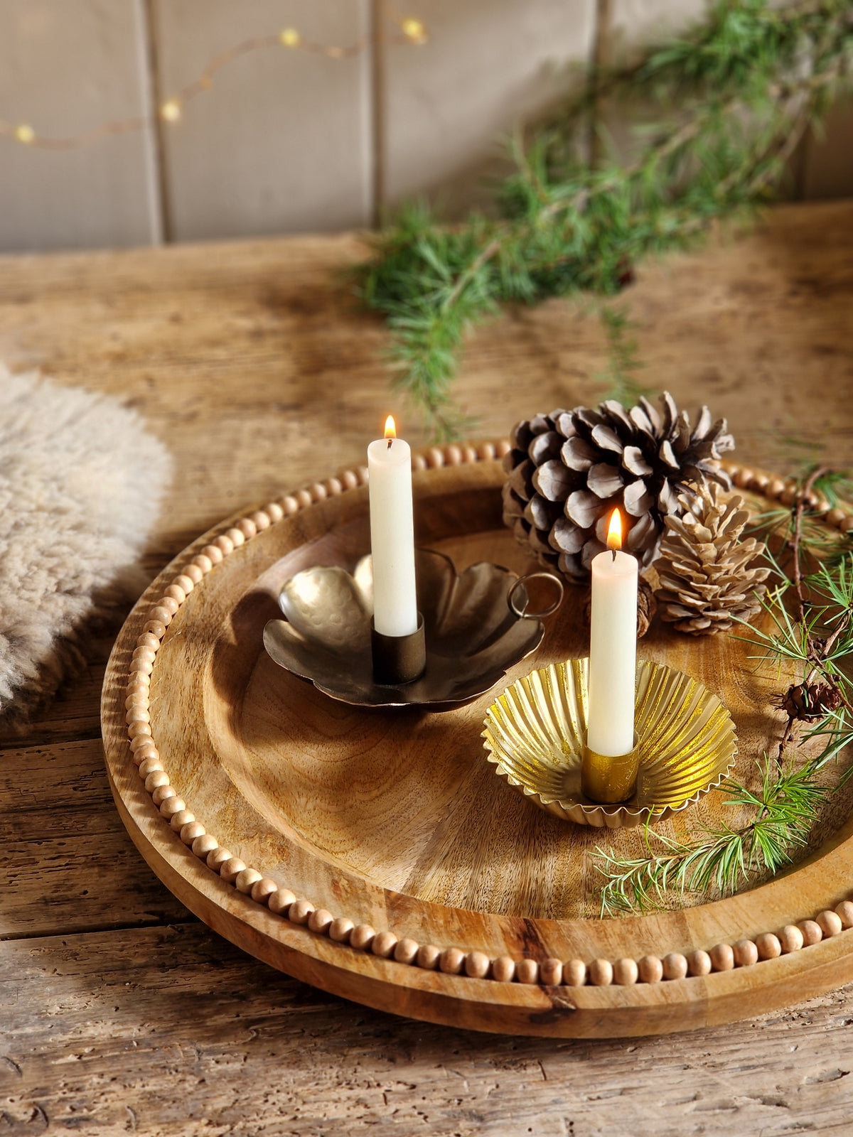 Round wooden wall-mounted candle holder with a beaded edge, holding two white taper candles, a pinecone, and a gold-toned shell ornament, styled against a rustic wood wall.