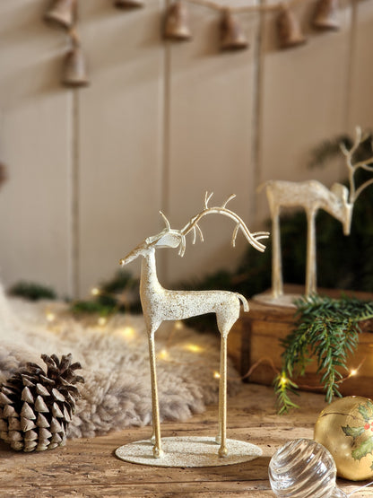 A decorative reindeer figurine with antlers and long legs, finished in a textured off-white and gold glittery material, standing on a round base on a rustic wooden surface. The scene is styled with a pinecone, greenery, warm fairy lights, and another similar figurine in the background.