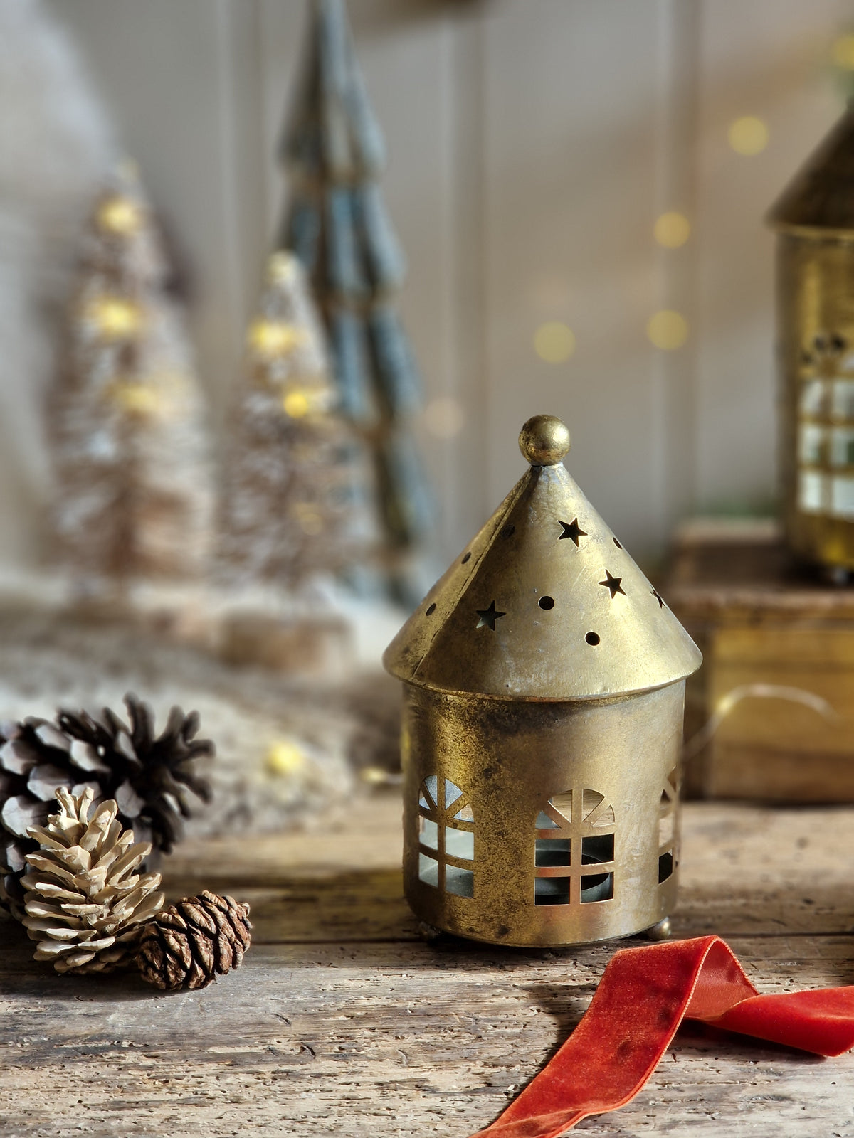 A small gold-toned metal Christmas house lantern with star and window cutouts sits on a rustic wooden surface, surrounded by pinecones and a red ribbon, with softly blurred holiday decorations in the background.
