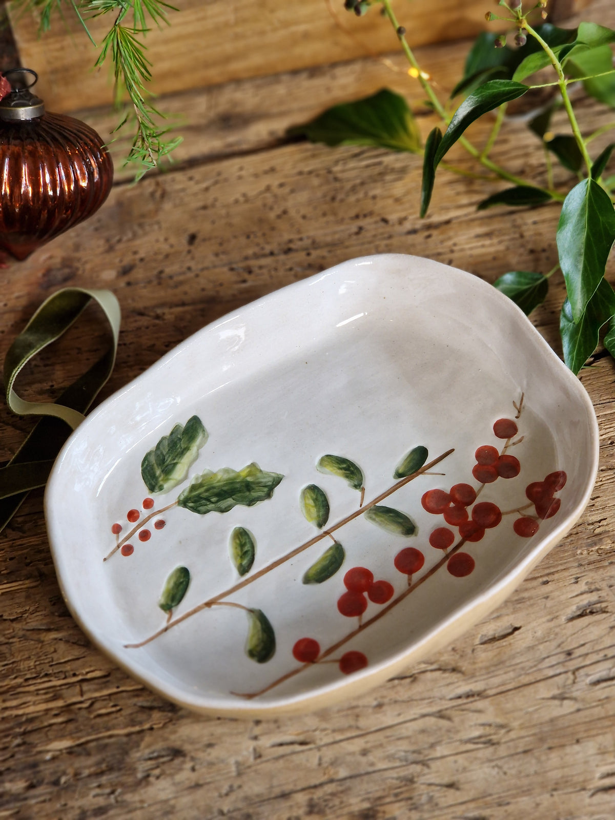 An oval white ceramic dish with a hand-painted holly design featuring green leaves and red berries, displayed on a rustic wooden surface with green foliage and a copper ornament nearby.