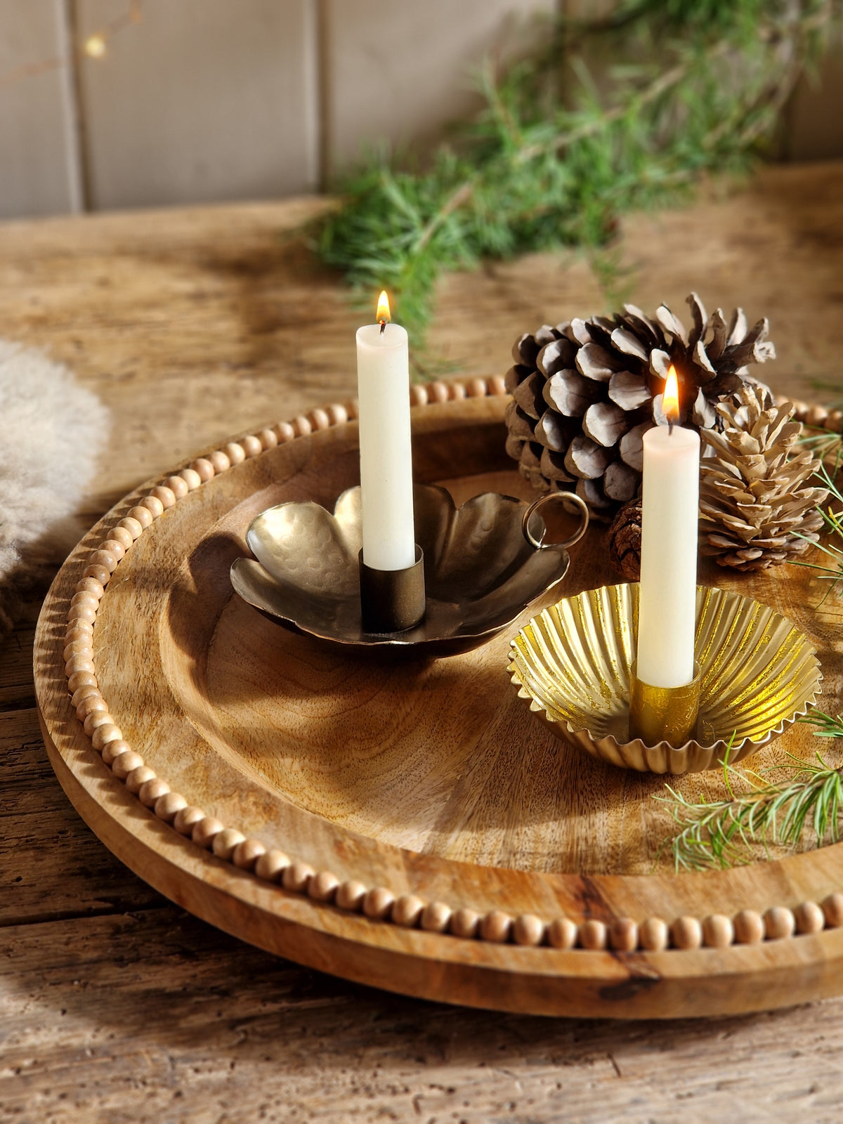 A decorative round wooden wall-mounted holder with carved beaded edges, holding two white taper candles in metallic holders, alongside a pinecone and greenery, against a rustic wood background.