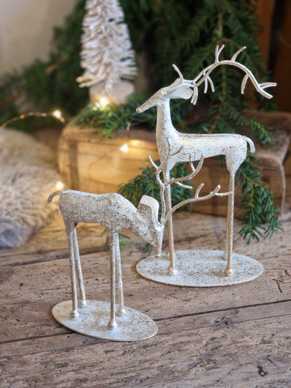 Two distressed white reindeer figurines with antlers and branch-like legs, displayed on oval bases on a rustic wooden surface with a blurred Christmas tree and warm lights in the background.