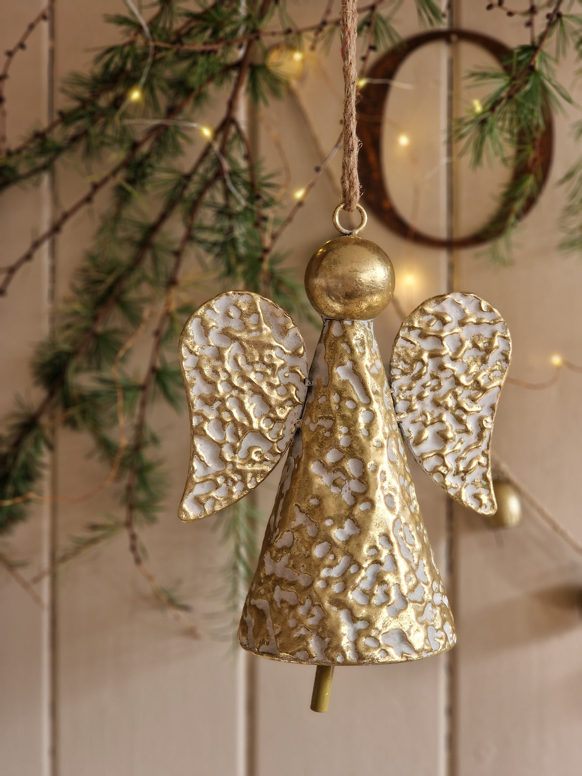 A gold and white textured angel Christmas ornament hanging from a twine string against a light wood paneled background with pine branches and warm string lights.