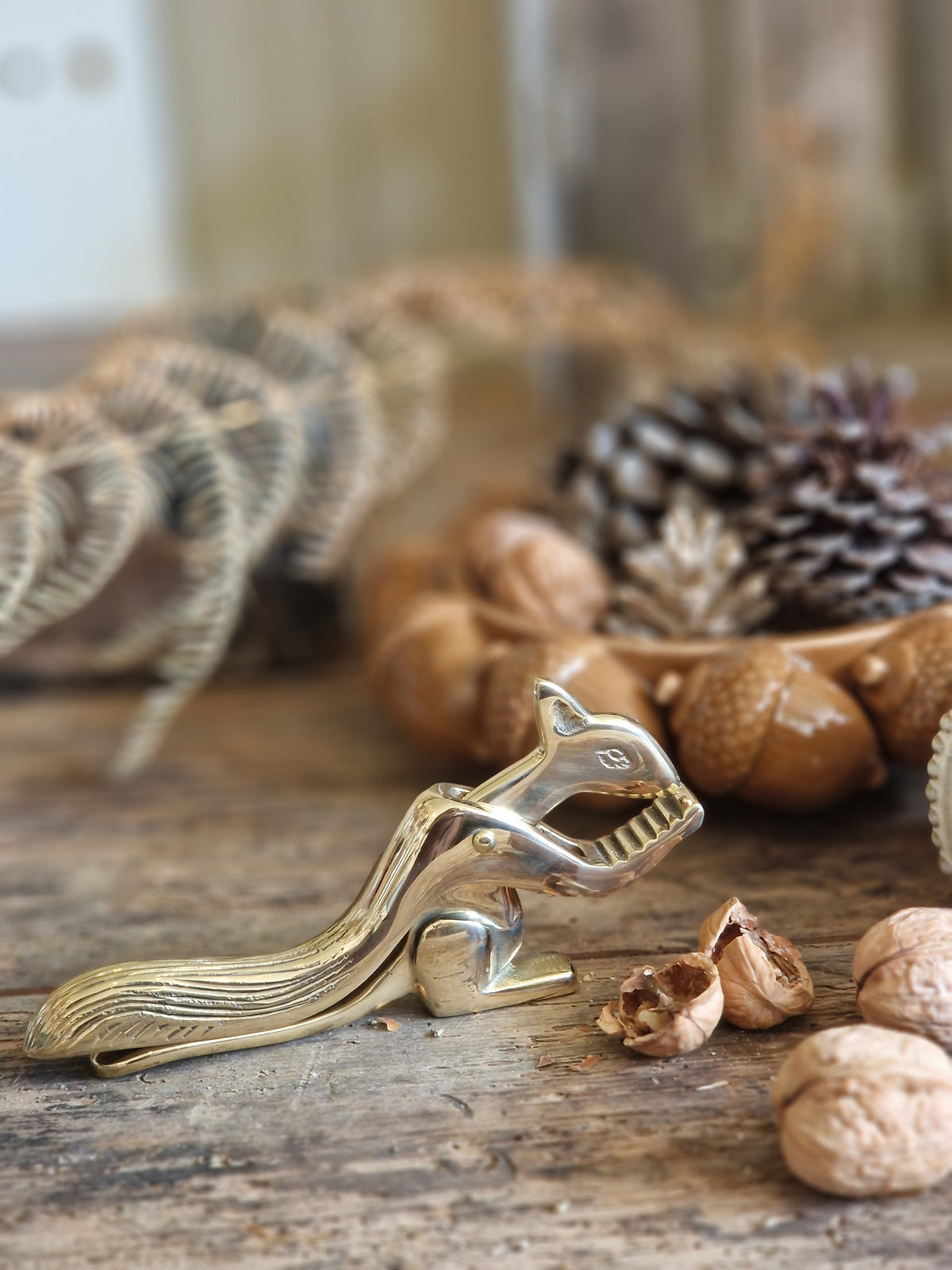 A polished brass squirrel-shaped nut cracker resting on a rustic wooden table, surrounded by walnuts, acorns, and pinecones.