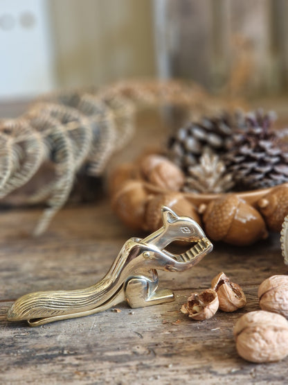 A polished brass squirrel-shaped nut cracker resting on a rustic wooden table, surrounded by walnuts, acorns, and pinecones.