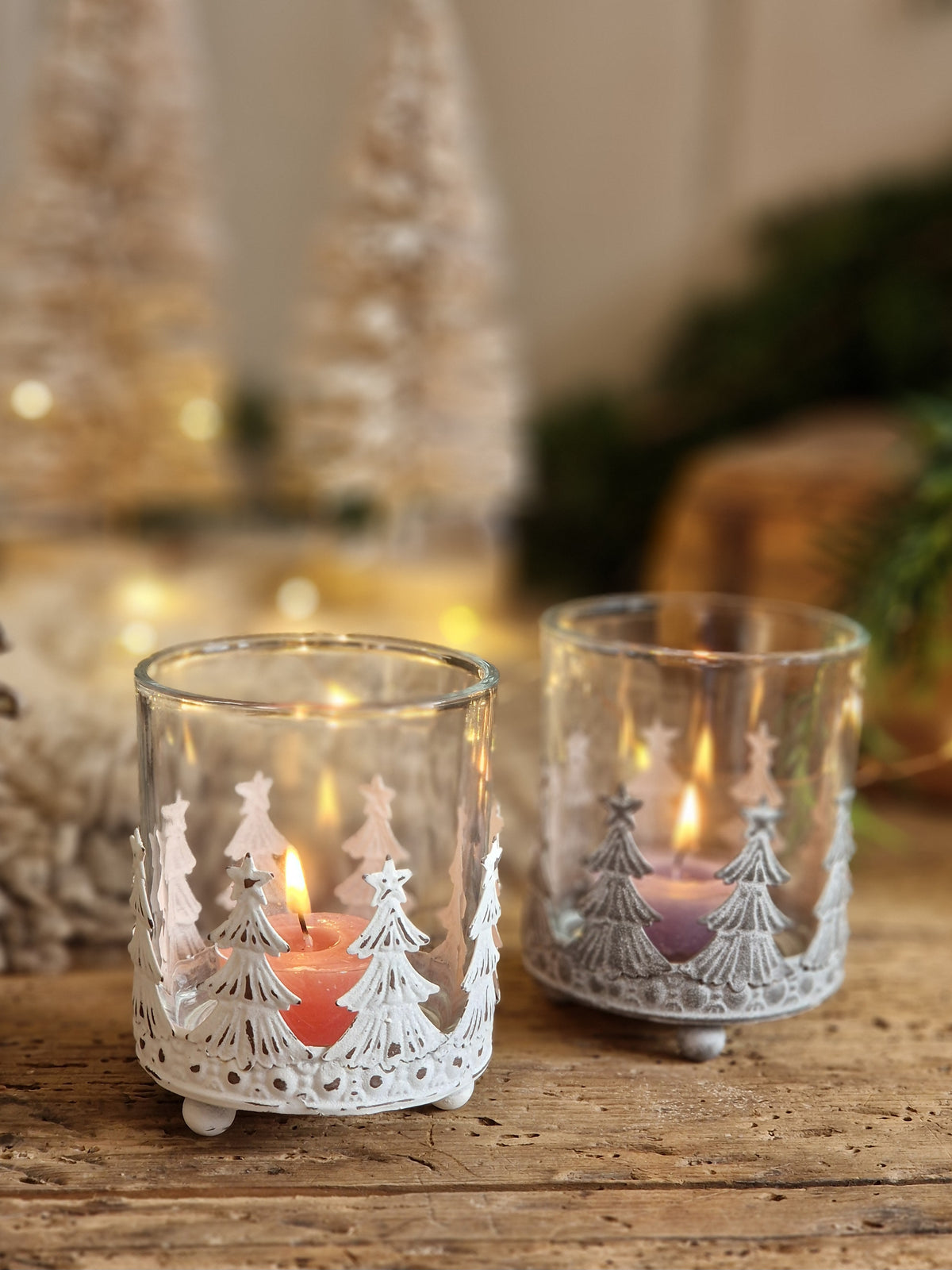 Two clear glass candle holders with white metal tree cutouts, each containing a small lit tealight candle, displayed on a rustic wooden surface with a softly blurred holiday background.
