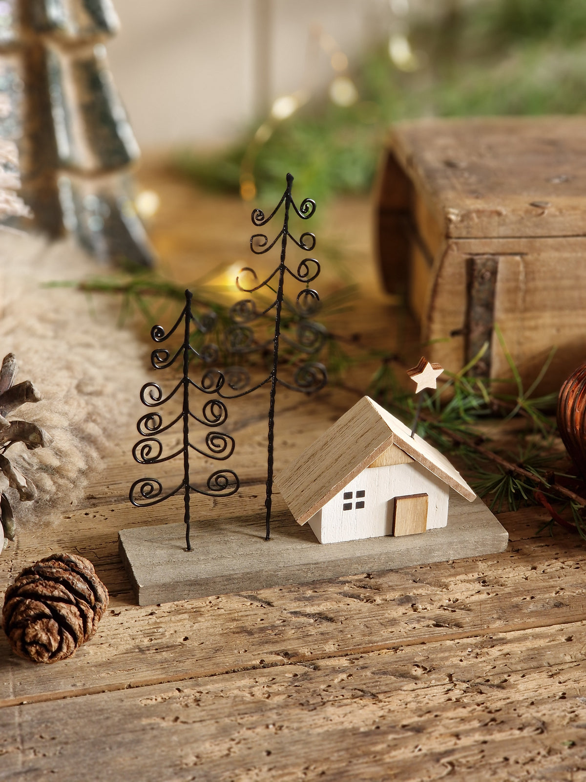 A miniature holiday cottage figurine with a light wood roof and white body, set on a gray base next to two black metal wire pine trees, styled on a rustic wooden surface with pinecones and greenery.