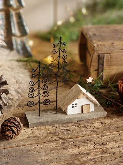 A miniature holiday cottage figurine with a light wood roof and white body, set on a gray base next to two black metal wire pine trees, styled on a rustic wooden surface with pinecones and greenery.