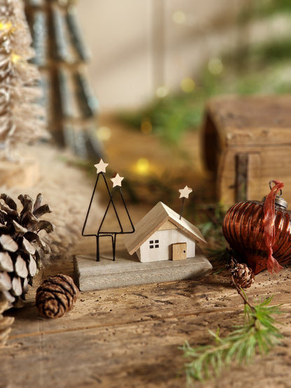 A close-up of rustic holiday decorations on a weathered wooden surface, including a small white house ornament, pinecones, evergreen branches, and a copper-colored ornament, with soft warm bokeh lights in the background.
