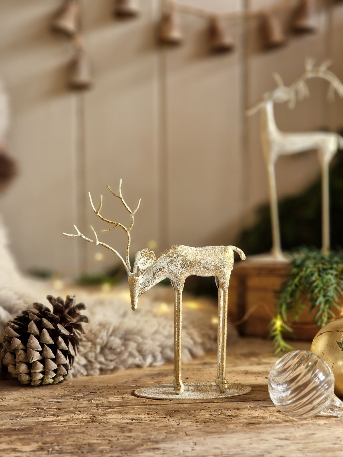 A small decorative reindeer figurine with a glittery gold finish stands on a rustic wooden surface, surrounded by holiday decor including a pinecone, a glass ornament, and another blurred reindeer statue in the background.