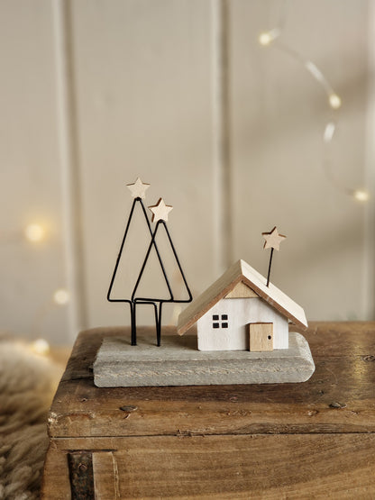 A rustic holiday wall decoration featuring a small white wooden house with a gray stone-textured base, mounted on a wooden panel with wire tree silhouettes and star accents.