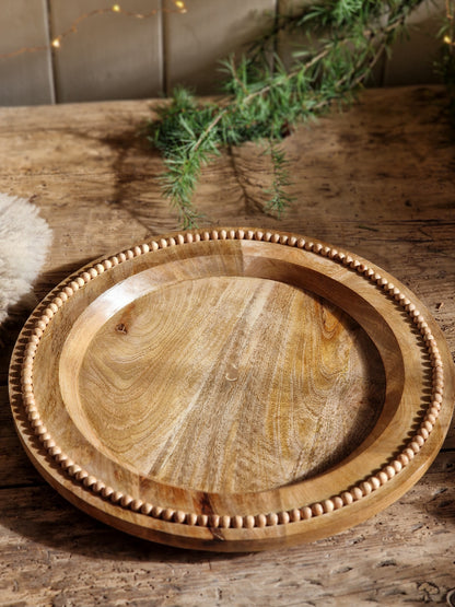 A round wooden serving tray with a decorative beaded rim, displayed on a rustic wooden table with pine branches and warm string lights in the background.