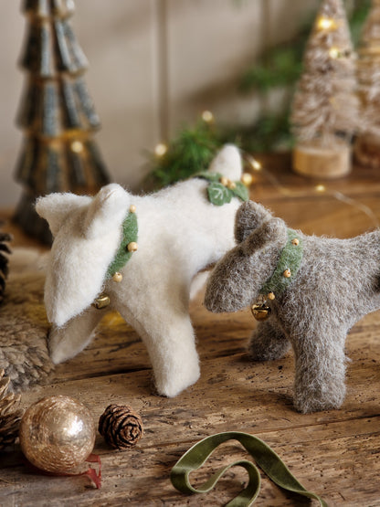 Two felted wool dog ornaments, one cream-white and one gray, wearing green leaf collars with small gold bells, posed on a rustic wooden surface with Christmas decorations in the background.