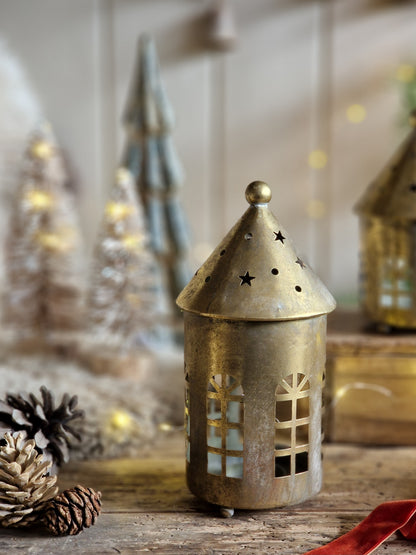 A small decorative brass lantern with a pointed star-topped roof and window cutouts stands on a rustic wooden surface. Pinecones and blurred holiday trees with warm lights appear in the background, creating a cozy festive setting.