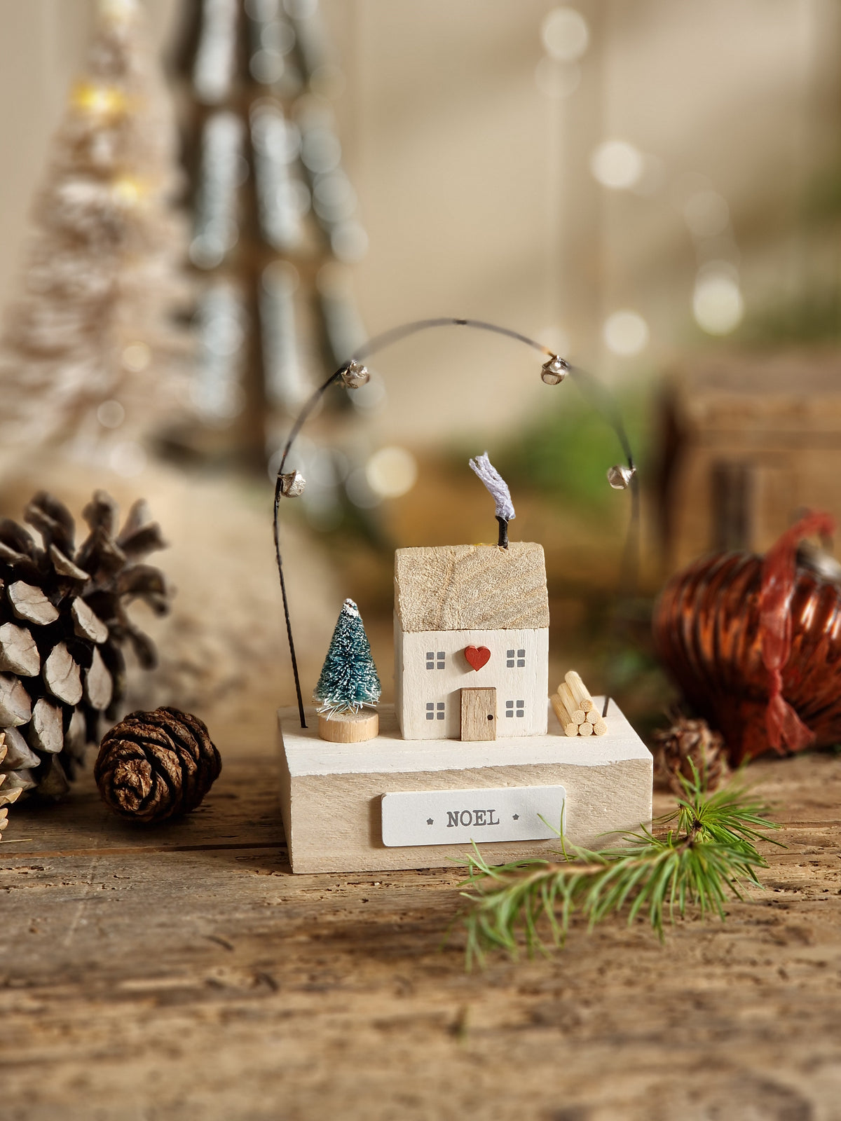A close-up of a rustic wooden house-shaped ornament or decor piece mounted on a wooden surface, surrounded by pinecones, greenery, and warm blurred bokeh lights.