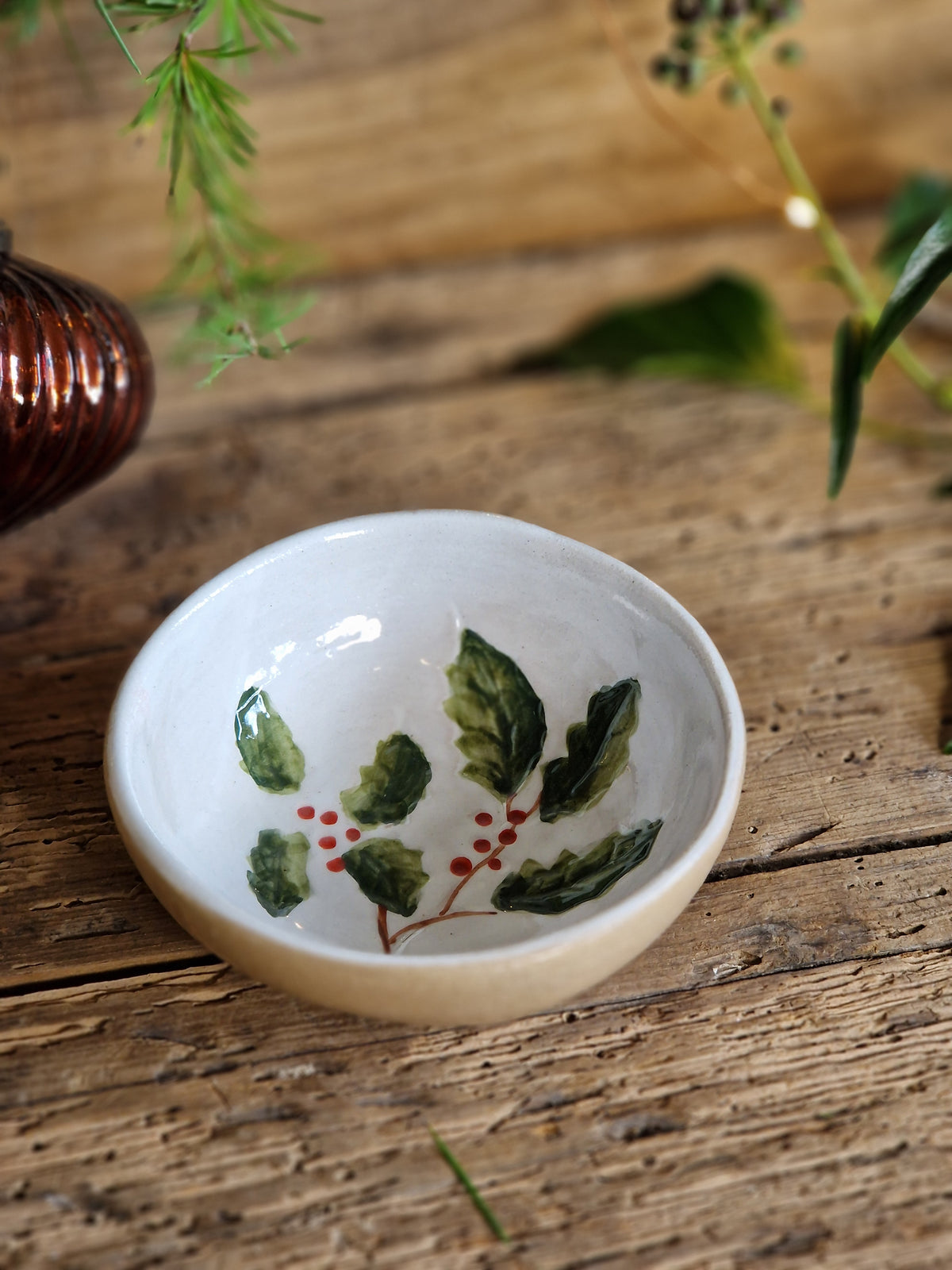 A small white ceramic bowl with a hand-painted green holly leaf and red berry design inside, resting on a rustic wooden surface.