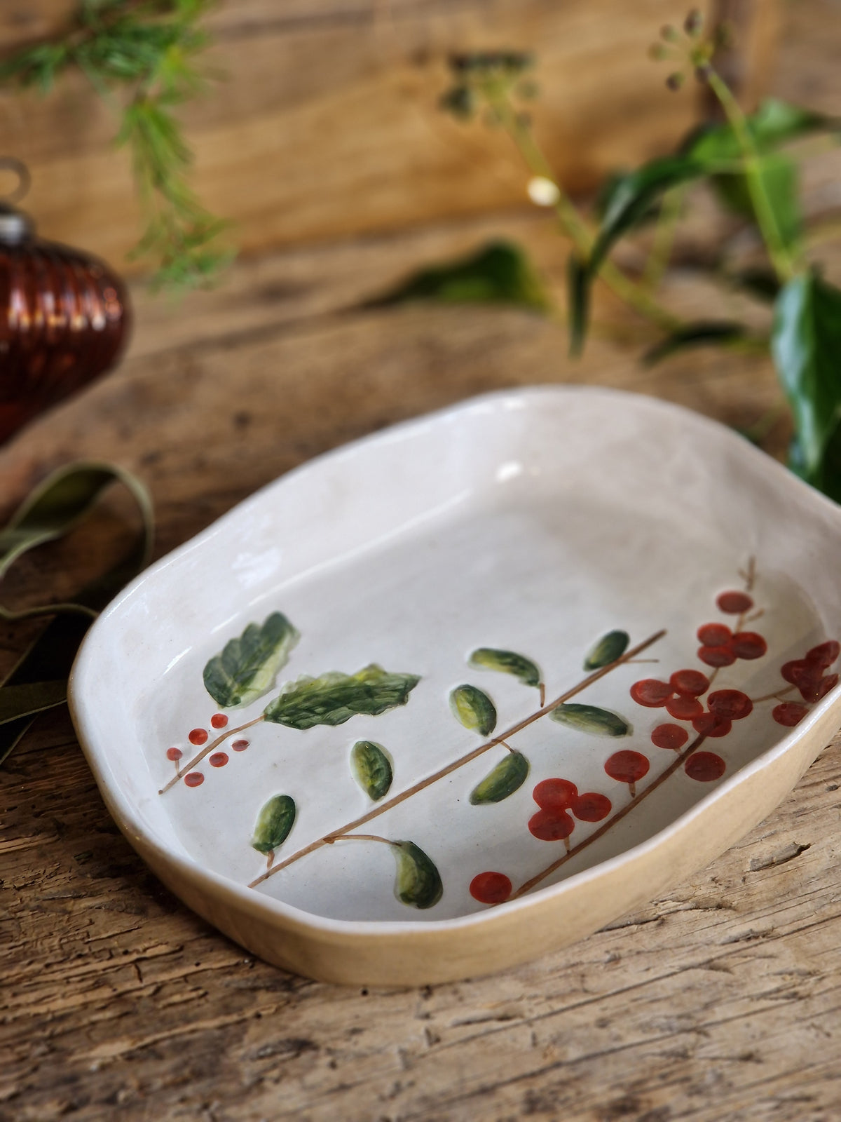 A hand-painted ceramic serving dish with a natural beige rim, featuring a green and red holly berry design on a rustic wooden background.