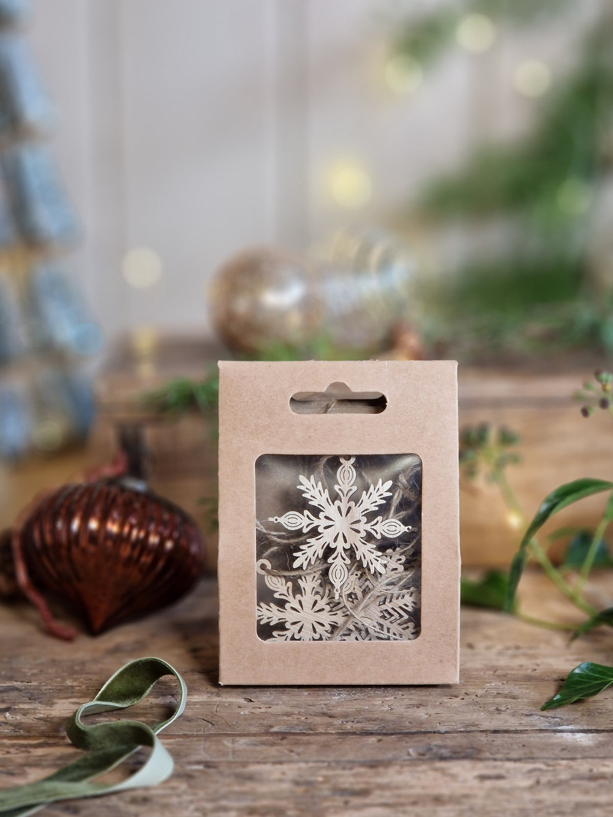 A small kraft paper hanging gift box with a clear window showing white snowflake ornaments inside, set on a rustic wooden surface with blurred holiday decor in the background.