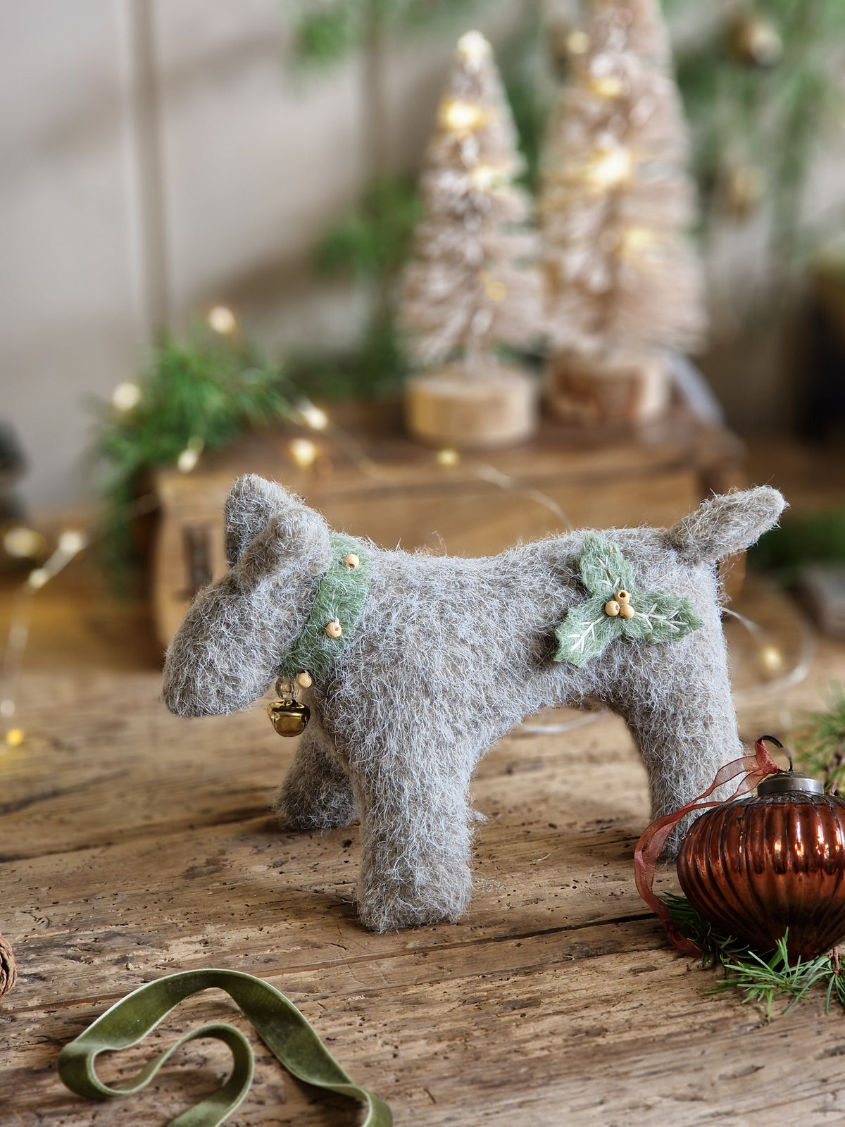 A handmade gray felt dog figurine decorated with a green holly leaf and gold bell, displayed on a rustic wooden surface with holiday decorations in the background.
