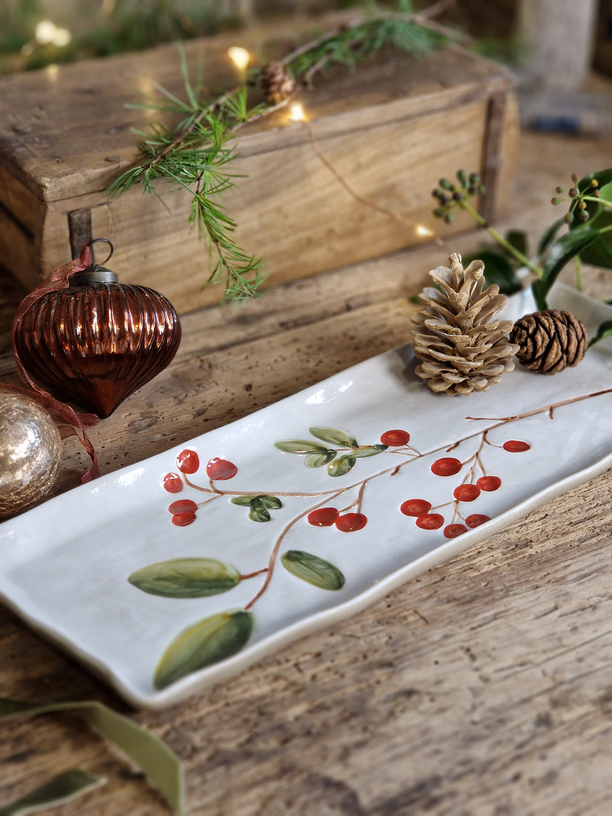 A rectangular white ceramic tray with a hand-painted winter berry branch design, styled on a rustic wooden surface with pinecones, evergreen sprigs, and warm fairy lights.