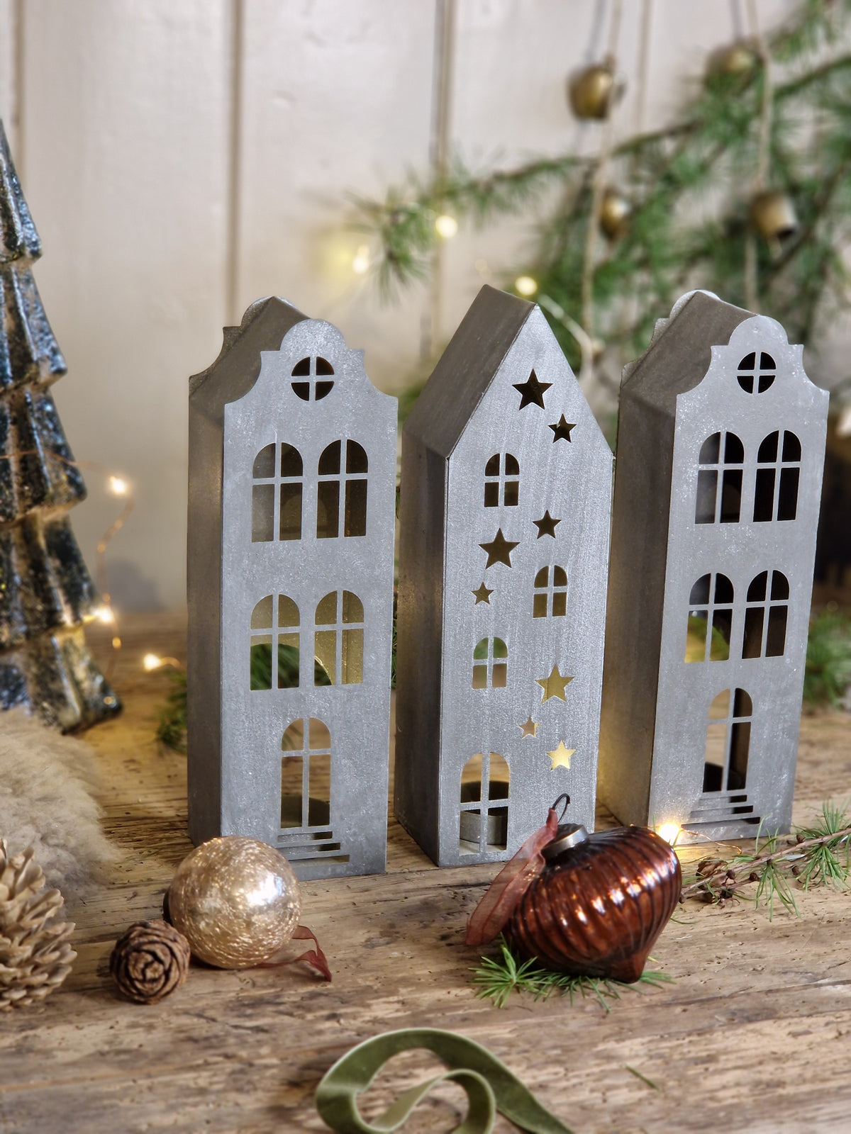 Three rustic metal Christmas house ornaments with star and window cutouts, displayed on a wooden surface with holiday decorations including pine branches, ornaments, and fairy lights.