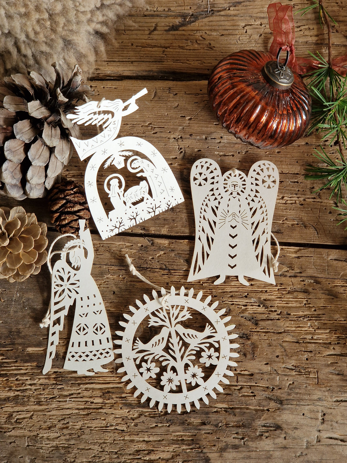 Four white laser-cut wooden Christmas ornaments with intricate folk-art designs, arranged on a rustic wooden surface alongside pinecones, a copper ornament, and evergreen branches.