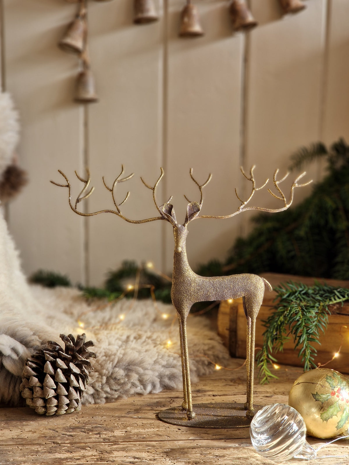 A decorative metallic reindeer figurine with antlers stands on a tabletop in a styled holiday setting, surrounded by a pinecone, a gold ornament, a glass ornament, greenery, and warm string lights.