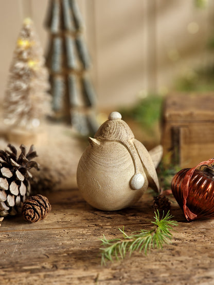 A decorative ceramic owl figurine in a natural beige tone, styled on a rustic wooden wall alongside pinecones, evergreen branches, and a red ornament.