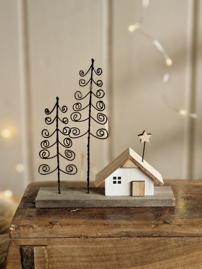 A small decorative holiday scene with two black metal spiral trees and a tiny white house with a light wood roof and star on a rustic wooden surface against a softly blurred indoor background.